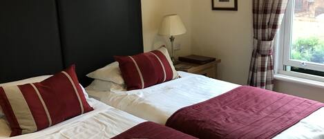 A double bed with white and red bedding, a remote control on the bed, a nightstand with a lamp, and a window with curtains.