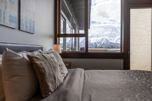 A bedroom with a bed, pillows, a nightstand, a lamp, and a window with a mountain view.