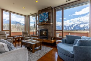 A living room with a fireplace, large windows, and a mountain view. - Historic 901 Penthouse with Incredible Views (Fernie)
