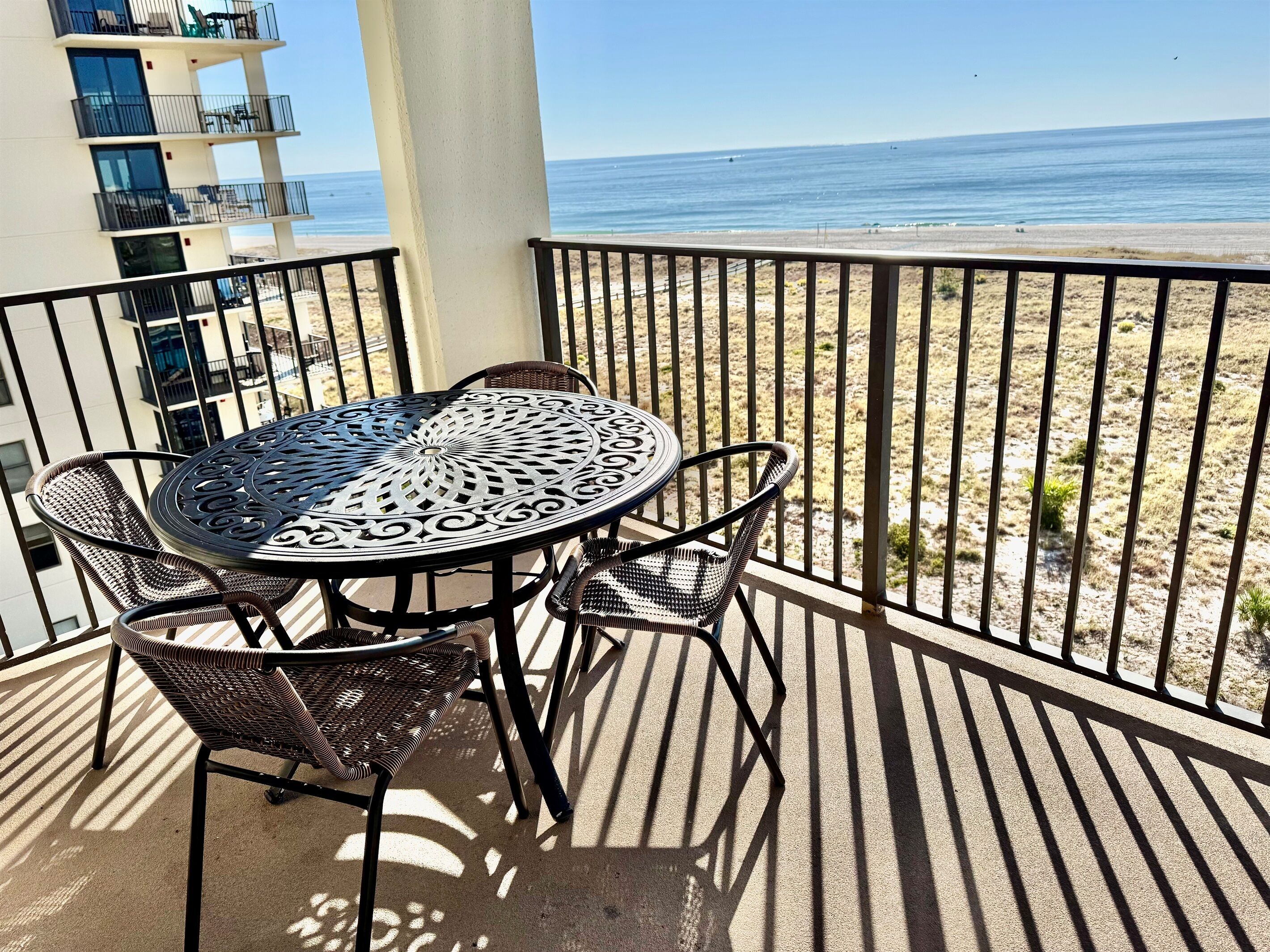 A balcony with a round metal table and wicker chairs, overlooking a beach.