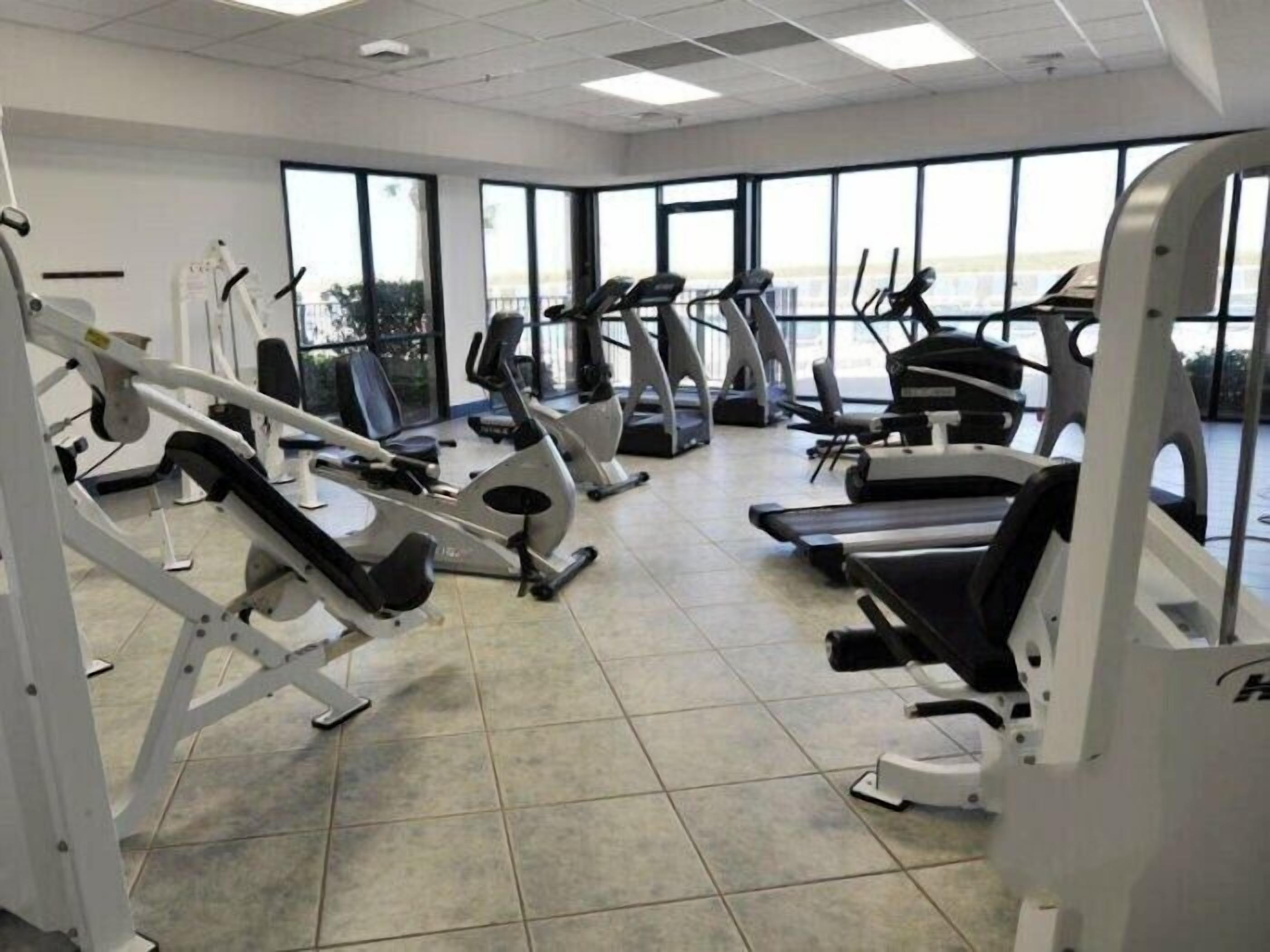Une salle de sport équipée de divers appareils d’exercice, dotée de grandes fenêtres et d’un sol carrelé.