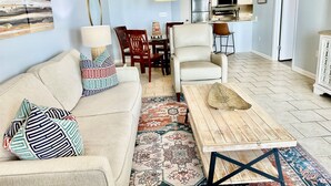 A living room with a sofa, patterned pillows, a coffee table, and a rug. There is a dining area with chairs and a table, and a kitchen with appliances and cabinets. - Newly Renovated kitchen in this 2BR Beachfront Condo on the 6th Floor! (Orange Beach)