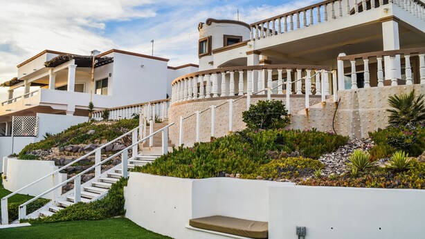 Un moderno edificio a più piani con facciata bianca, un balcone curvo e un giardino curato con diverse piante e un vialetto in pietra.