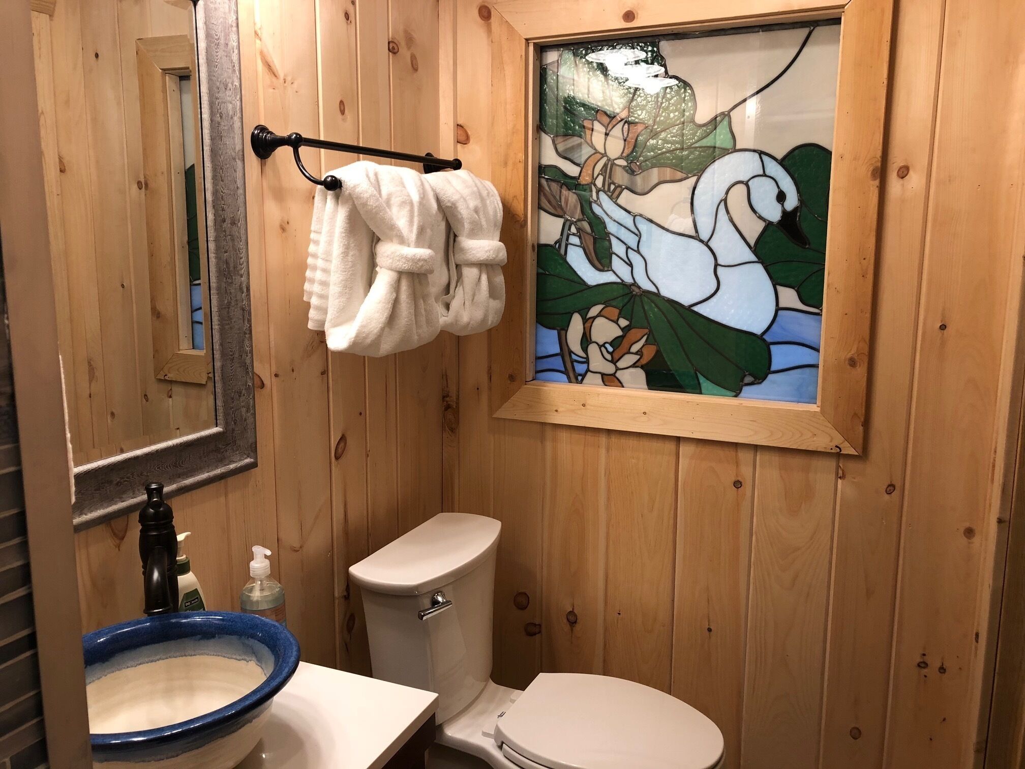 A wooden-paneled bathroom with a white toilet, a blue basin sink, a towel rack with white towels, and a stained glass window.