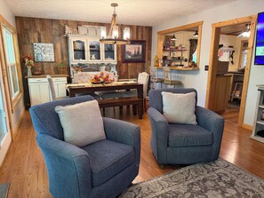 Een gezellige woonkamer met twee fauteuils, een eettafel en een keukenhoek met houten kasten en een kroonluchter.