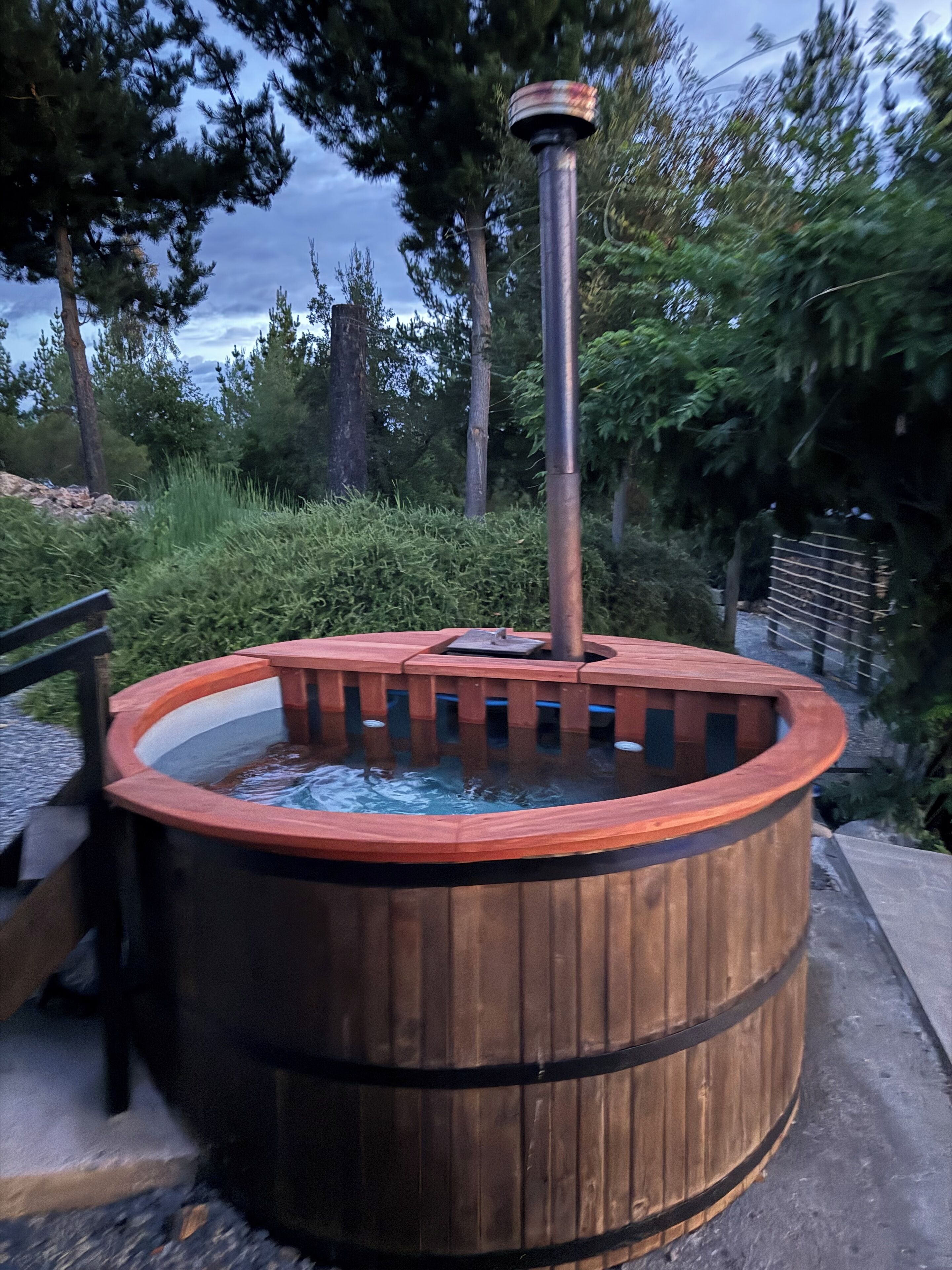 Un jacuzzi de madera con una chimenea metálica, rodeado de árboles y una terraza de madera.