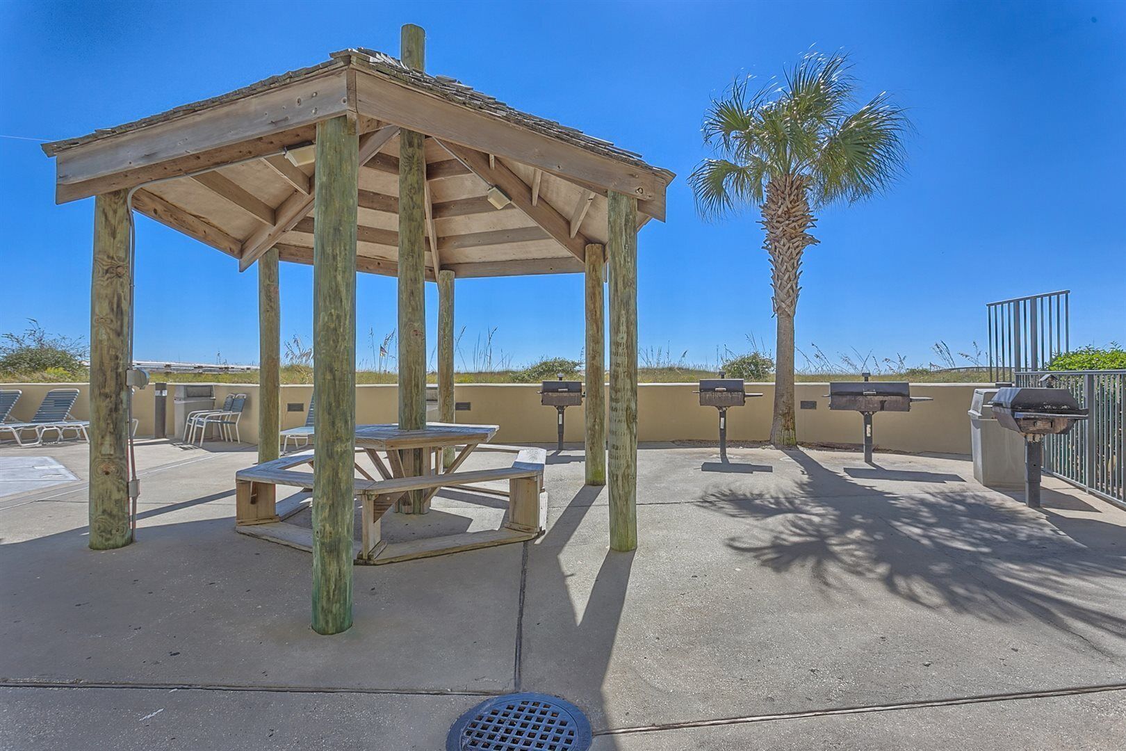 A wooden gazebo with picnic tables, a palm tree, and barbecue grills.