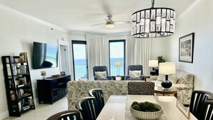Een moderne woonkamer met een houten eettafel, stoelen, een bank, een televisie, een boekenkast en uitzicht op de oceaan door grote ramen.