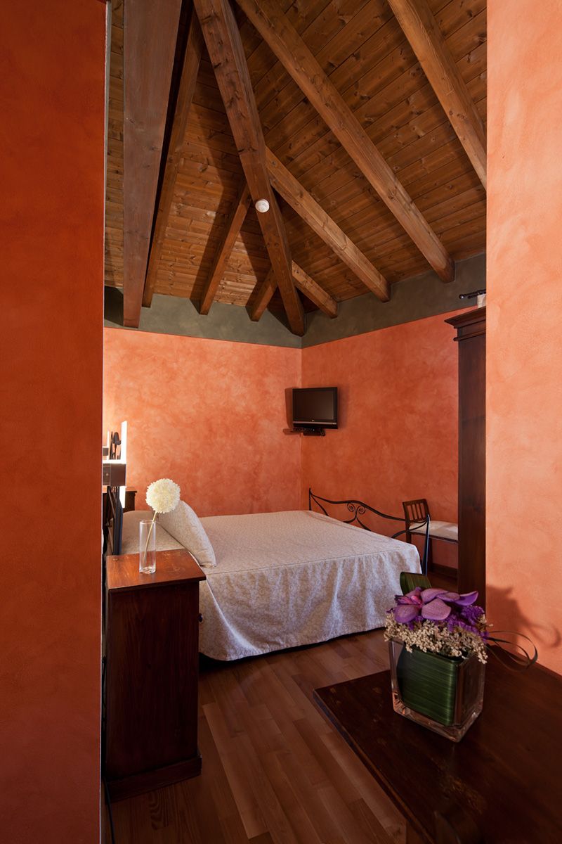 A bedroom with a wooden ceiling, a bed, a bedside table, a chair, and a TV mounted on the wall.