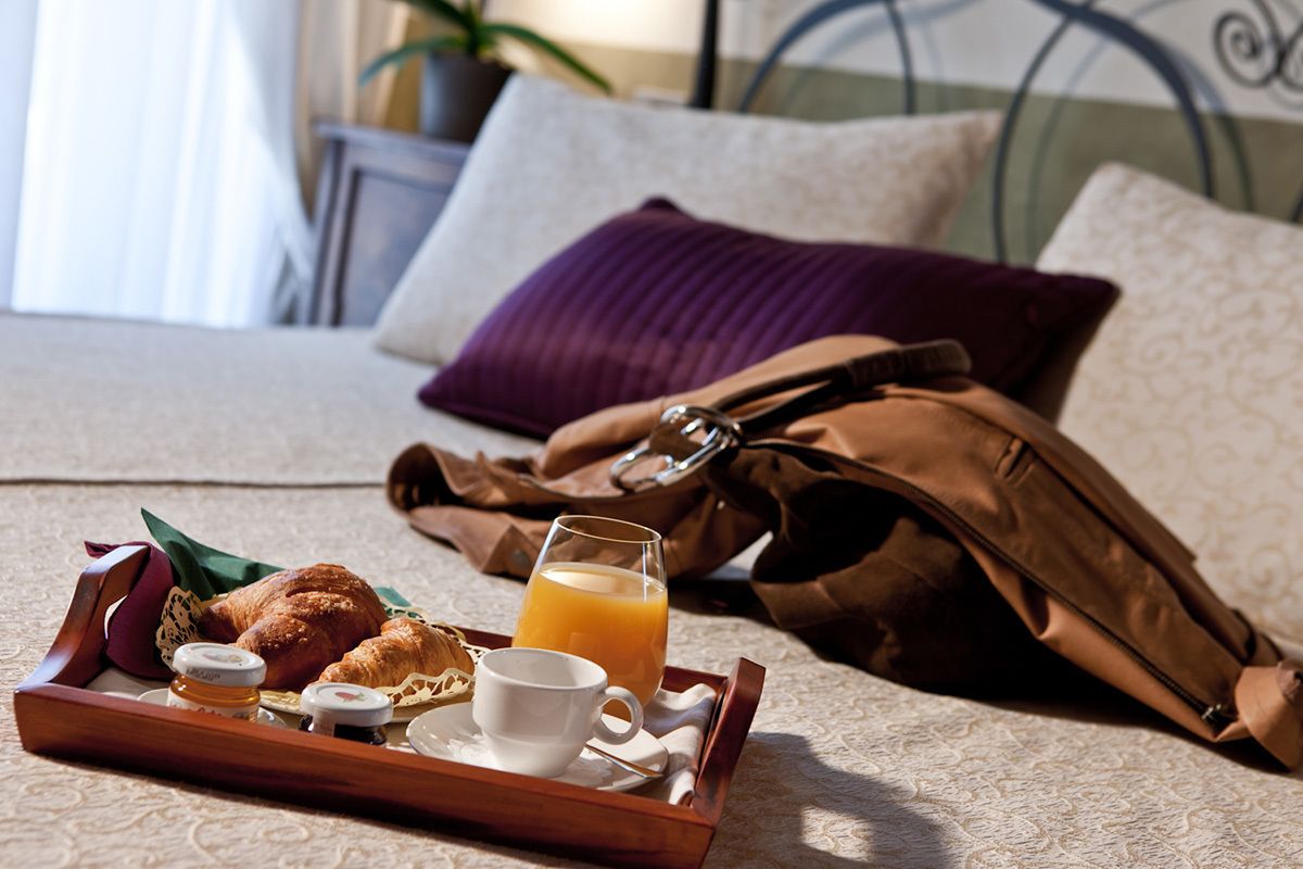 A breakfast tray with croissants, a cup, and orange juice on a bed with a brown bag and pillows.