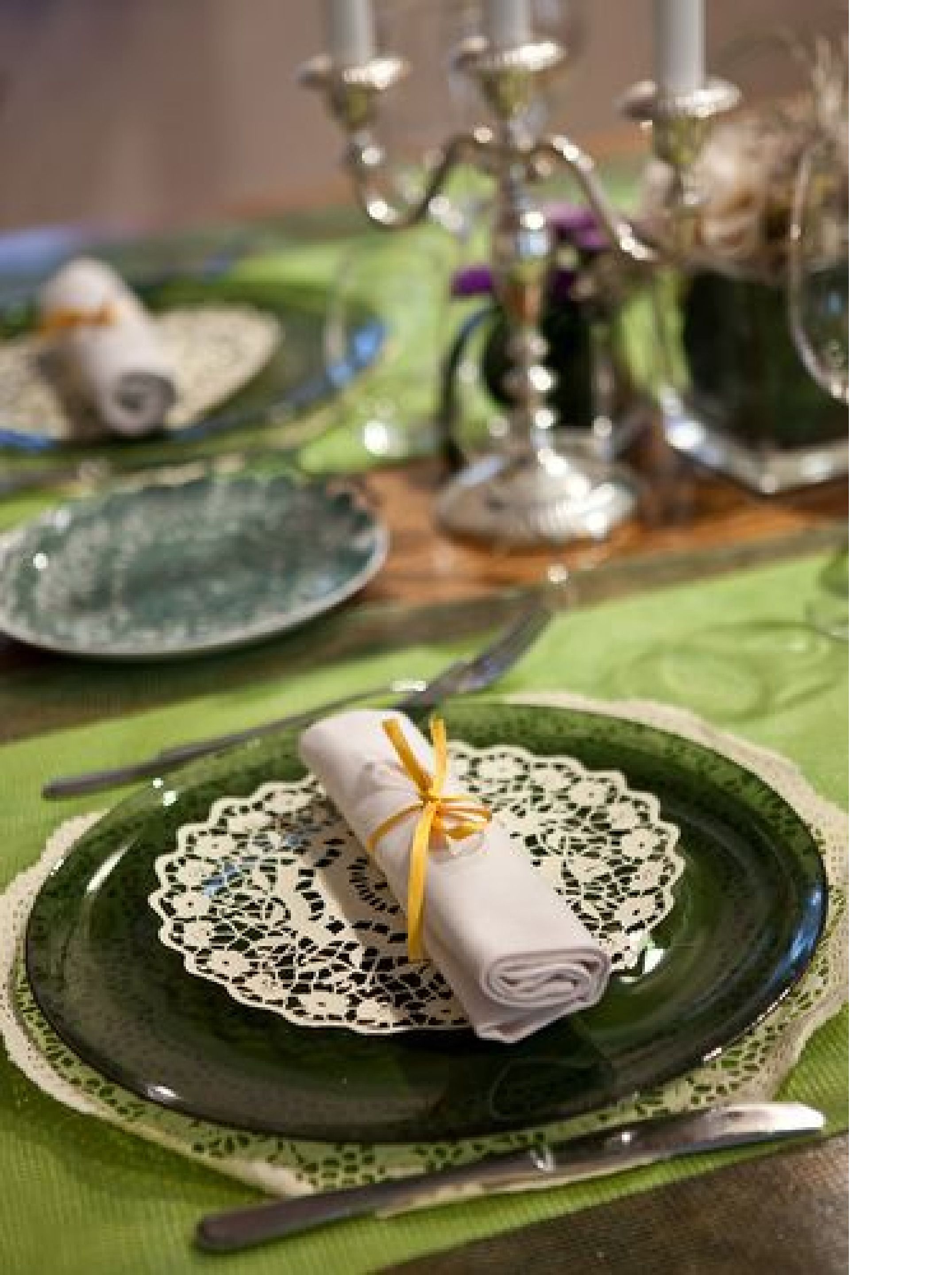 Una mesa dispuesta con platos verdes, un mantel de encaje, un servilleta doblada atada con un lazo amarillo y cubiertos.