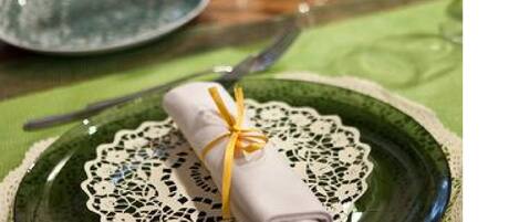 Una mesa dispuesta con platos verdes, un mantel de encaje, un servilleta doblada atada con un lazo amarillo y cubiertos.