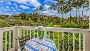 Balcón con mesa y sillas, con vista a un jardín tropical con palmeras y un cielo azul despejado.