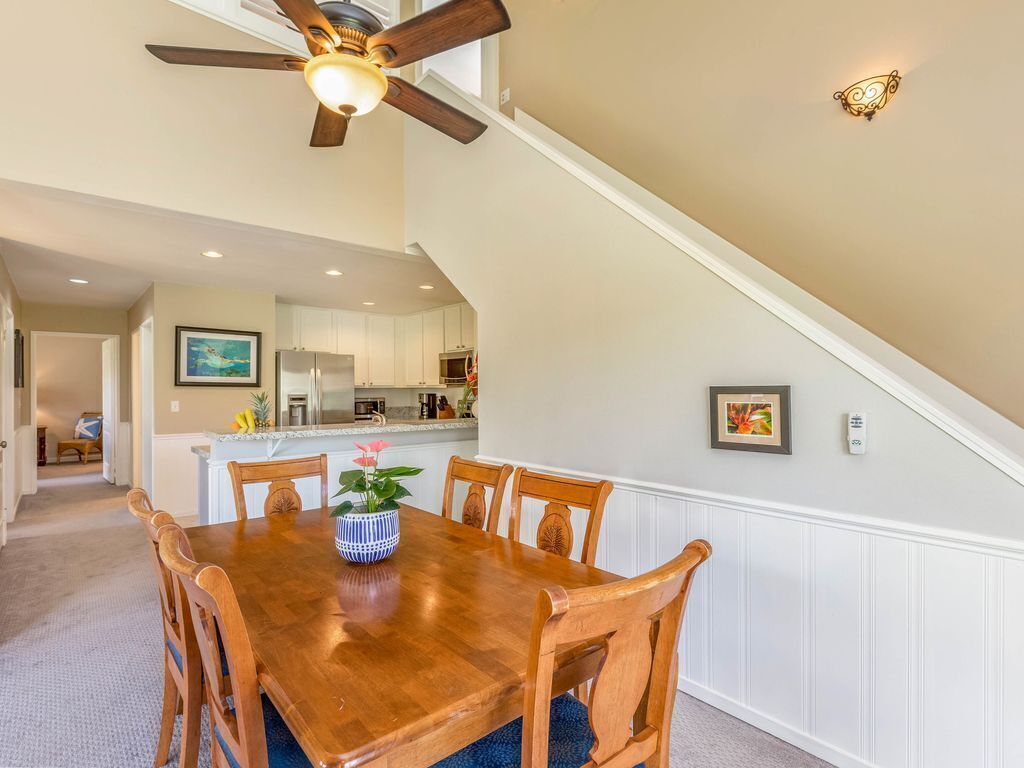 A dining area with a wooden table and chairs, a ceiling fan, and a kitchen in the background.
