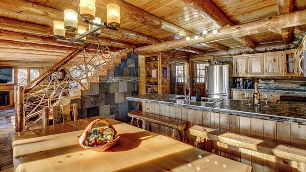 A rustic cabin interior with a stone fireplace, wooden beams, a wooden staircase, a kitchen with a large island, and a dining area with a basket of fruit.