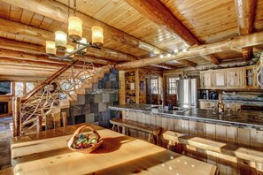 Interior rústico de una cabaña con chimenea de piedra, vigas de madera, una escalera de madera, una cocina con una isla grande y un comedor con una cesta de fruta.