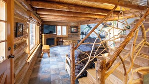A log cabin interior with a fireplace, wooden staircase, and a stone-tiled floor.