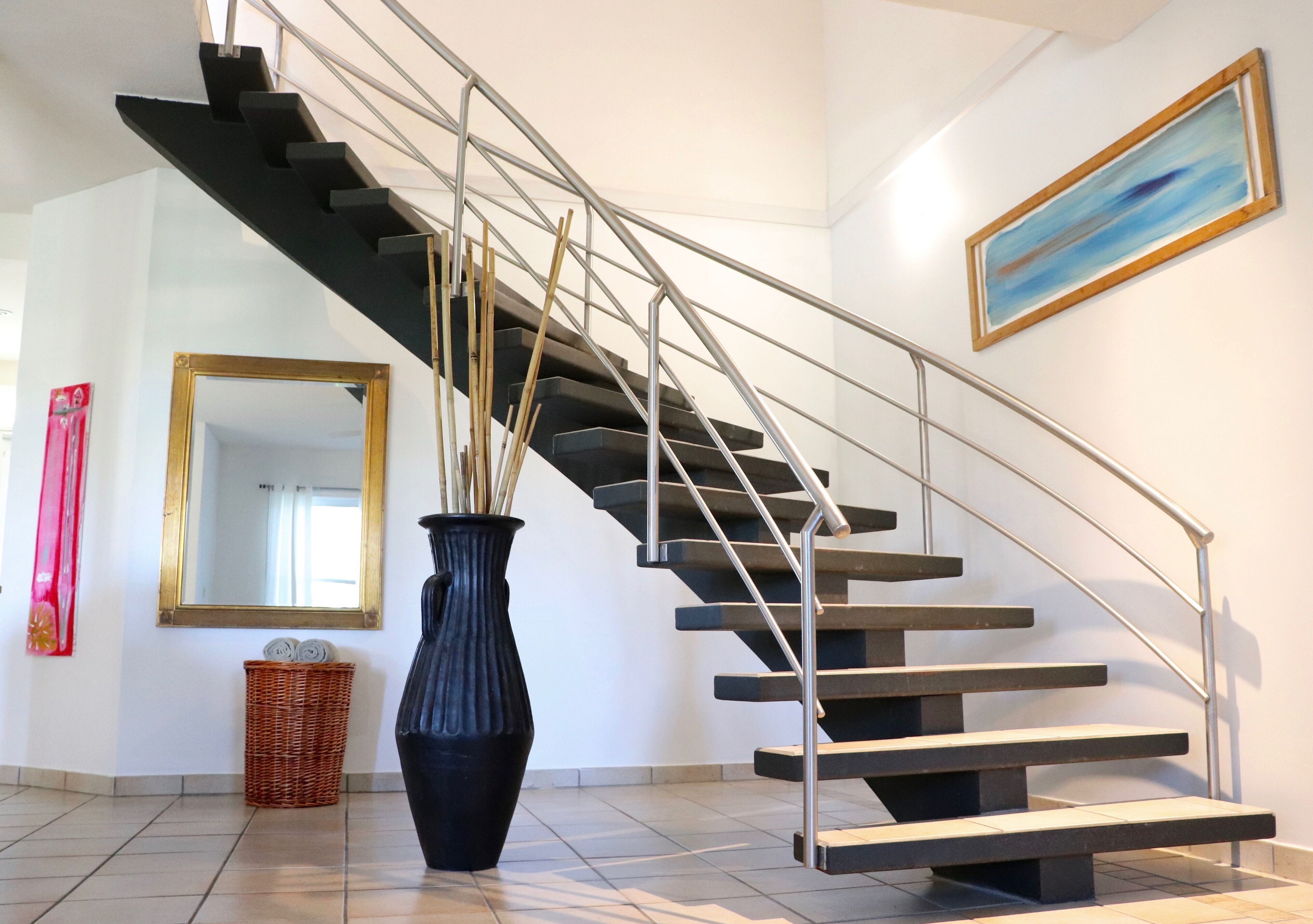 A modern staircase with metal railings, a large mirror, a vase with sticks, and a painting on the wall.