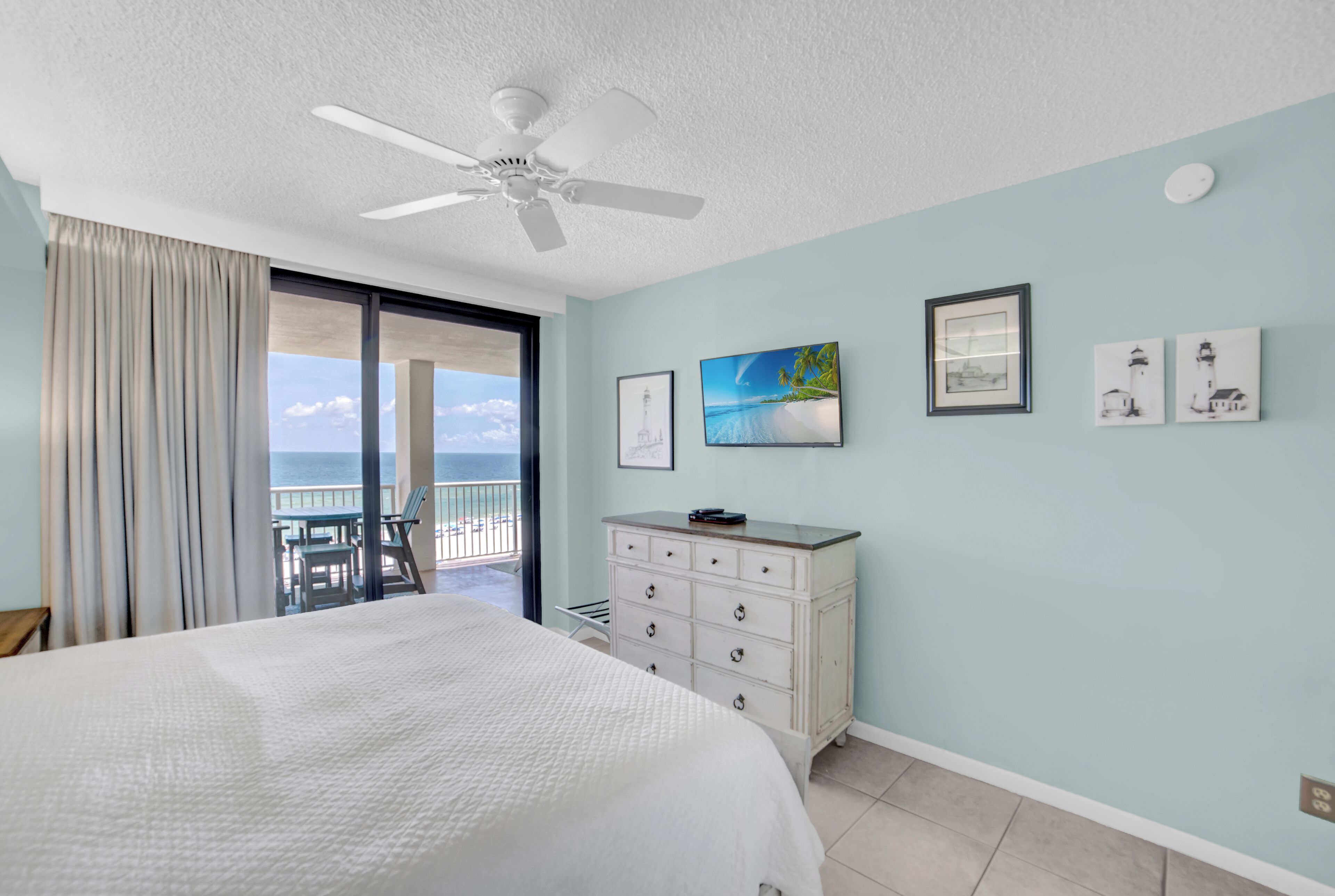 A bedroom with a bed, a dresser, and a ceiling fan. There are framed pictures on the wall and a view of the ocean through the window.