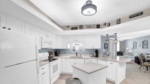 A modern kitchen with white cabinets, a central island, and a dining area with a table and chairs. - Summer House 704B by Liquid Life (Orange Beach)