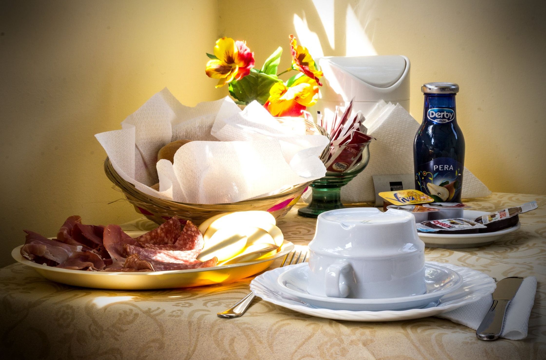 Une table mise pour le petit-déjeuner avec une assiette de charcuterie, un bol de pain, une tasse de café, une bouteille d’eau et un vase de fleurs.