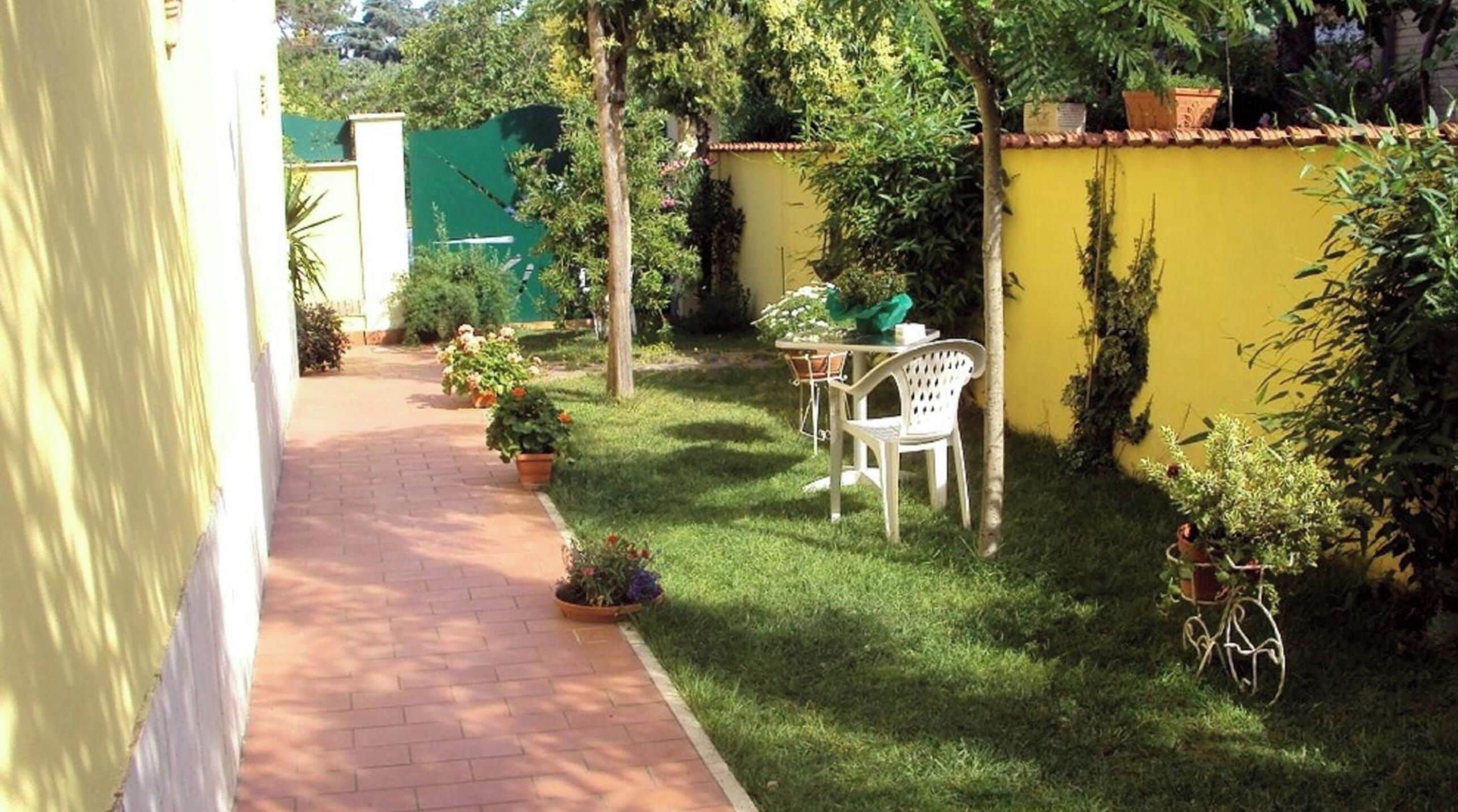 Un sendero pavimentado que conduce a una pequeña zona de patio con una mesa y sillas, rodeada de plantas en macetas y una pared amarilla.
