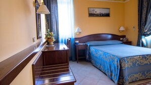 Habitación de hotel con escritorio de madera, cama con cubrecama azul y dorado, mesita de noche con lámpara y ventana con cortinas.