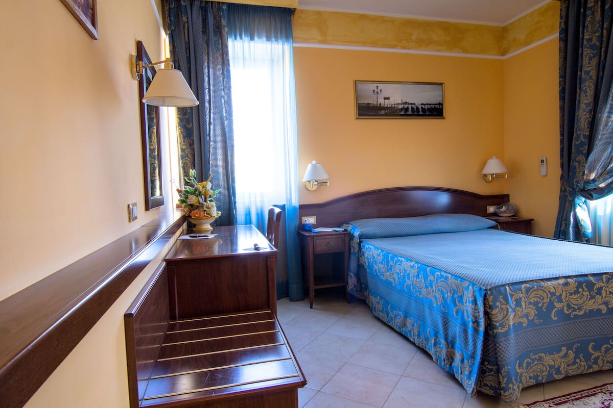 Habitación de hotel con escritorio de madera, cama con cubrecama azul y dorado, mesita de noche con lámpara y ventana con cortinas.