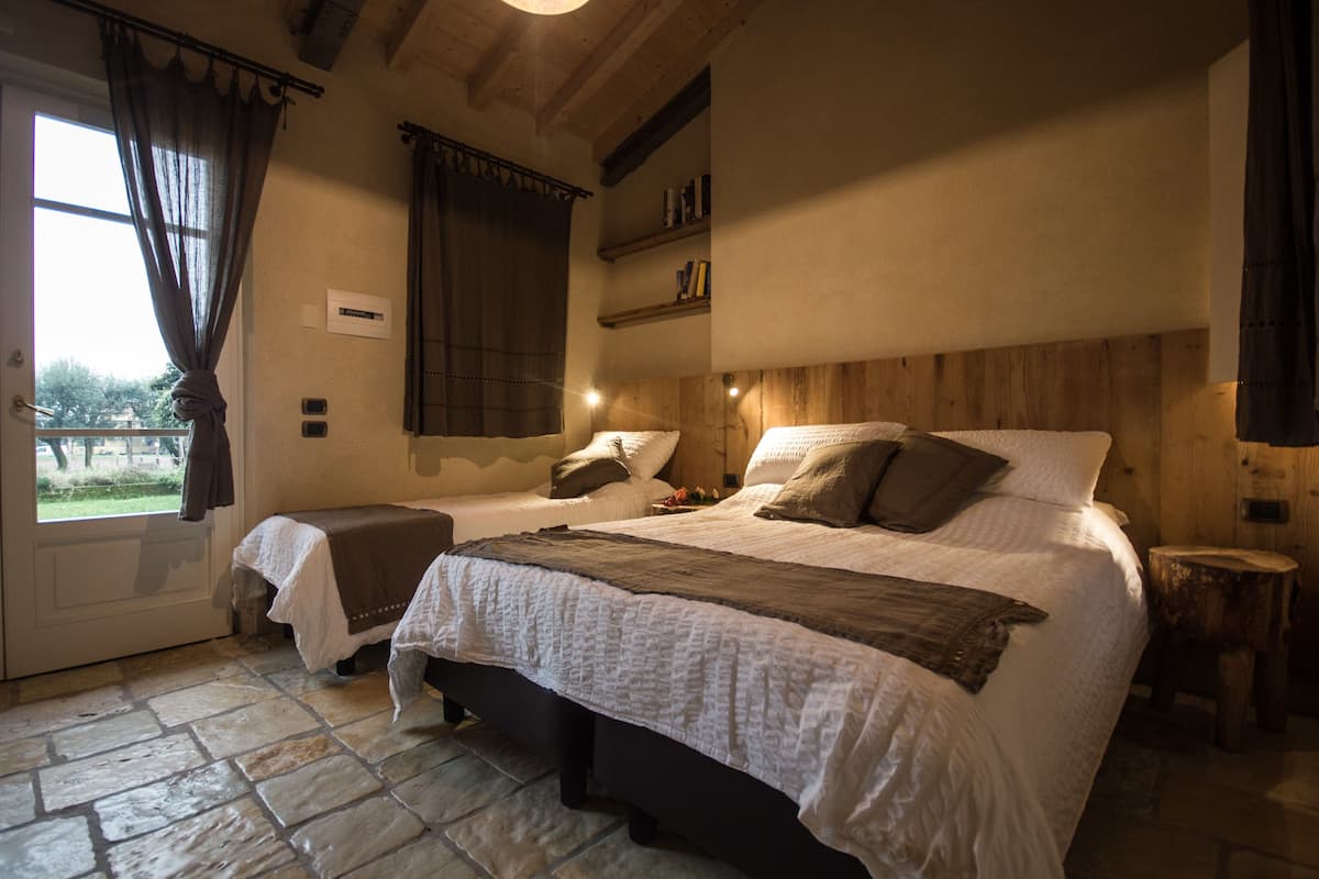 A bedroom with two beds, a stone floor, wooden headboard, and a window with curtains.