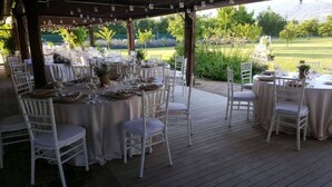 An outdoor dining area with round tables and white chairs, set up on a wooden deck with a view of a landscaped garden and distant hills. - Olimagio (Pietrasanta)