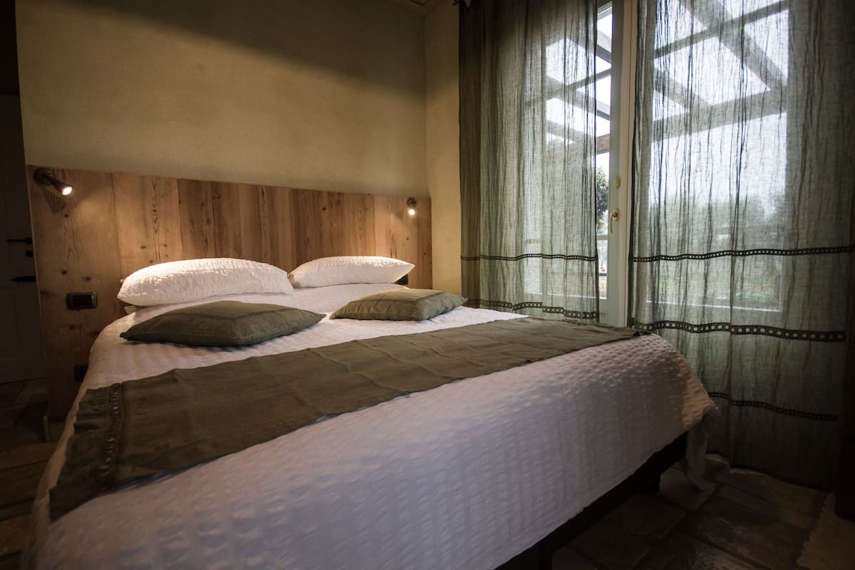A bedroom with a wooden headboard, a bed with white bedding, two pillows, and a view through sheer curtains.
