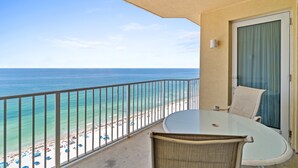 A balcony with a glass table and chairs overlooking a beach with umbrellas and the ocean.