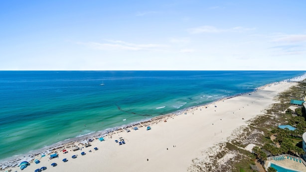 An aerial view of a beach with clear turquoise waters, a sandy shore, and a few umbrellas and beach chairs.