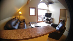 A living room with a brown leather sofa, a glass coffee table, and a TV stand with a flat-screen TV. - Beautiful Estes Park 2 Bedroom Loft in the ❤️ of town with Air Conditioning (Estes Park)