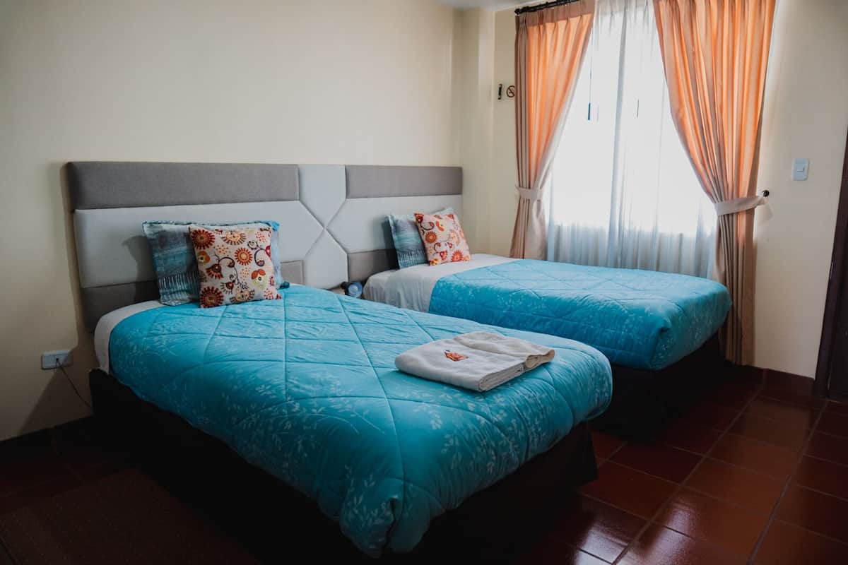 A hotel room with two beds, each with a blue quilted bedspread and decorative pillows. The room has a tiled floor and a window with orange curtains.