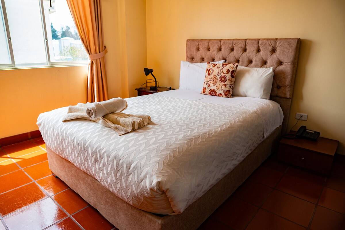 A neatly made bed with a patterned bedspread and two pillows, a bedside table with a lamp, and a window with curtains.