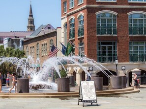 A public fountain with water jets, surrounded by brick buildings and flags, with a sign indicating no lifeguard on duty.
