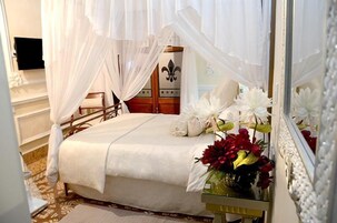 Een slaapkamer met een bed, witte gordijnen, een spiegel en een vaas met bloemen.