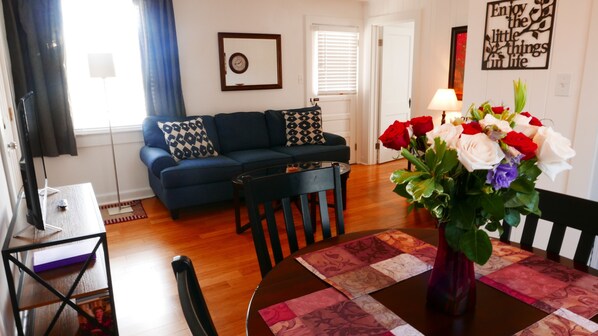 A living room with a sofa, a coffee table, and a dining table with chairs. There is a vase with flowers on the dining table.