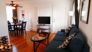 A living room with a sofa, a coffee table, a TV on a stand, a dining table with chairs, and a ceiling fan.