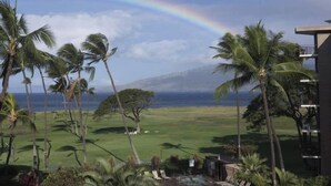 A view of a golf course with a rainbow, palm trees, and a building with a balcony. - Oceanfront penthouse with amazing views at Kauhale Makai (Kihei)