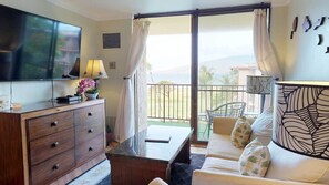 A modern living room with a flat-screen TV, a wooden cabinet, a sofa with patterned cushions, and a balcony with a view of the outdoors.
