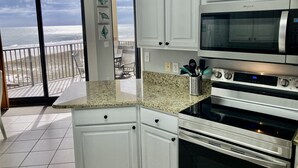 A kitchen with white cabinets, a granite countertop, a microwave, and an oven. There is a view of the ocean through a sliding glass door.