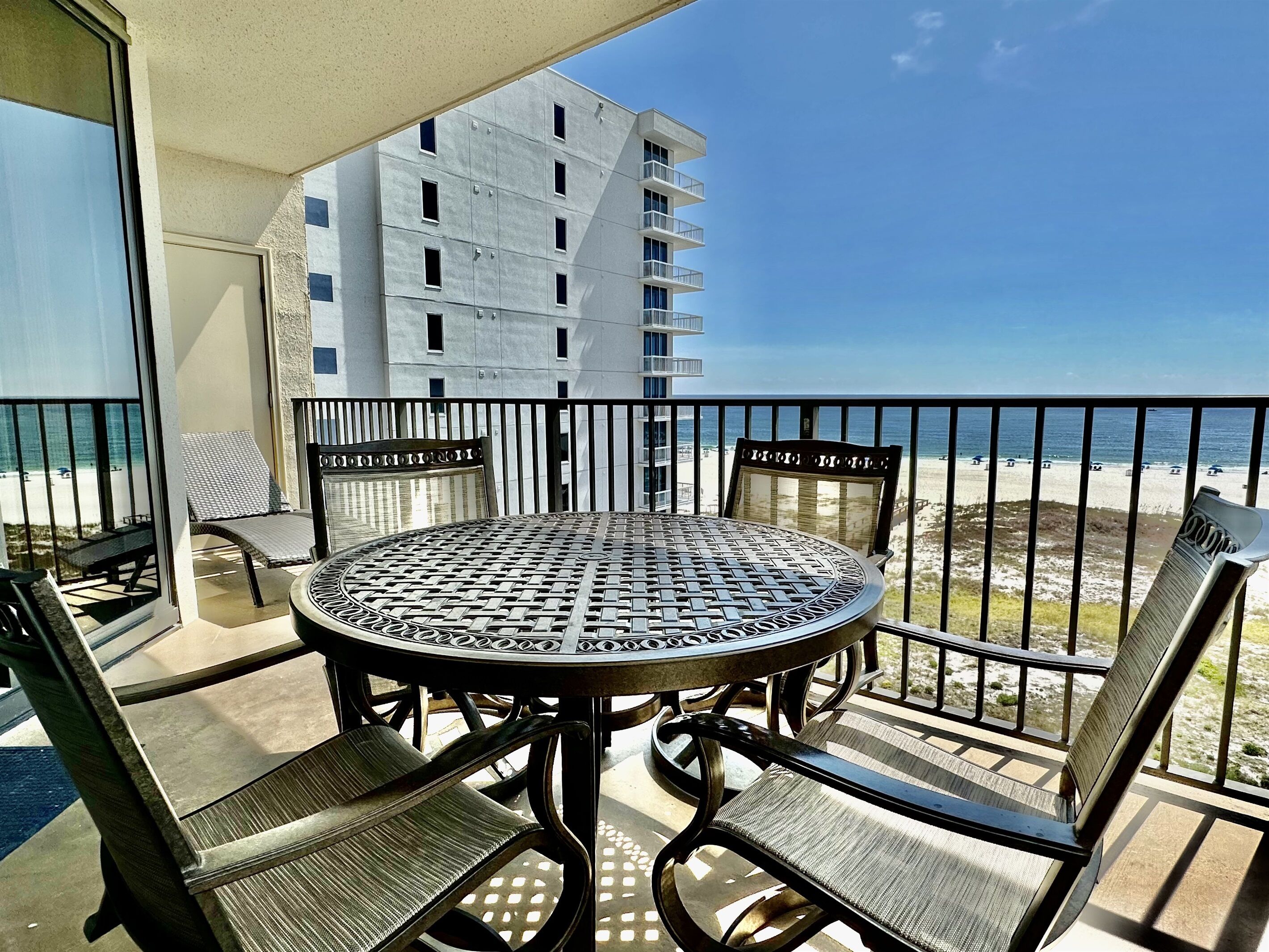 Un balcón con una mesa redonda y cuatro sillas, con vista a una playa y un edificio de apartamentos.