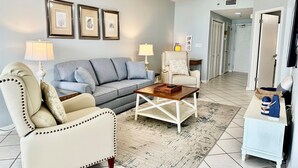 A living room with a grey sofa, a white armchair, a coffee table, and a ceiling fan. There are framed pictures on the wall and a lamp on the side table.