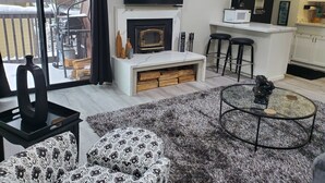 Un salon moderne avec une cheminée, un téléviseur fixé au-dessus, et un coin cuisine agrémenté d’une pancarte sur laquelle on peut lire : « Si vous voulez prendre votre petit-déjeuner au lit, dormez dans la cuisine ».