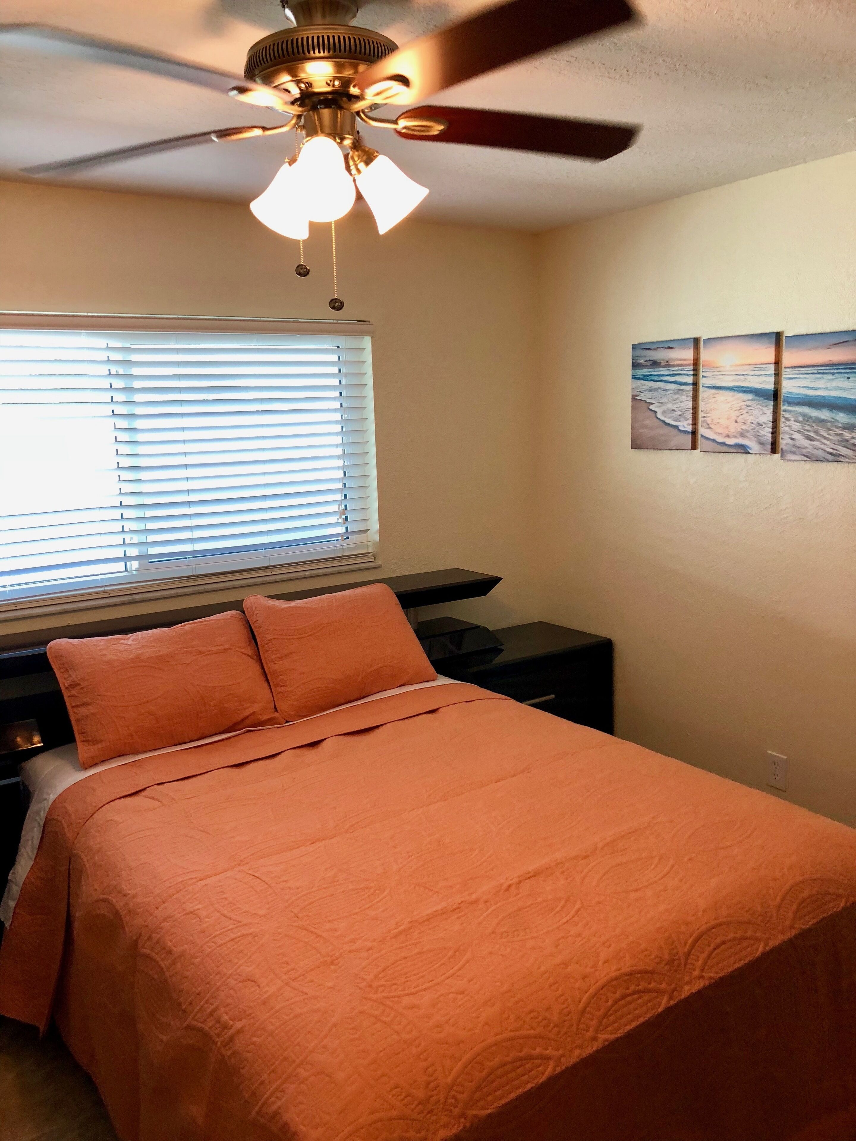 Un dormitorio con cama, ventilador de techo, ventana con persianas y un cuadro colgado en la pared.