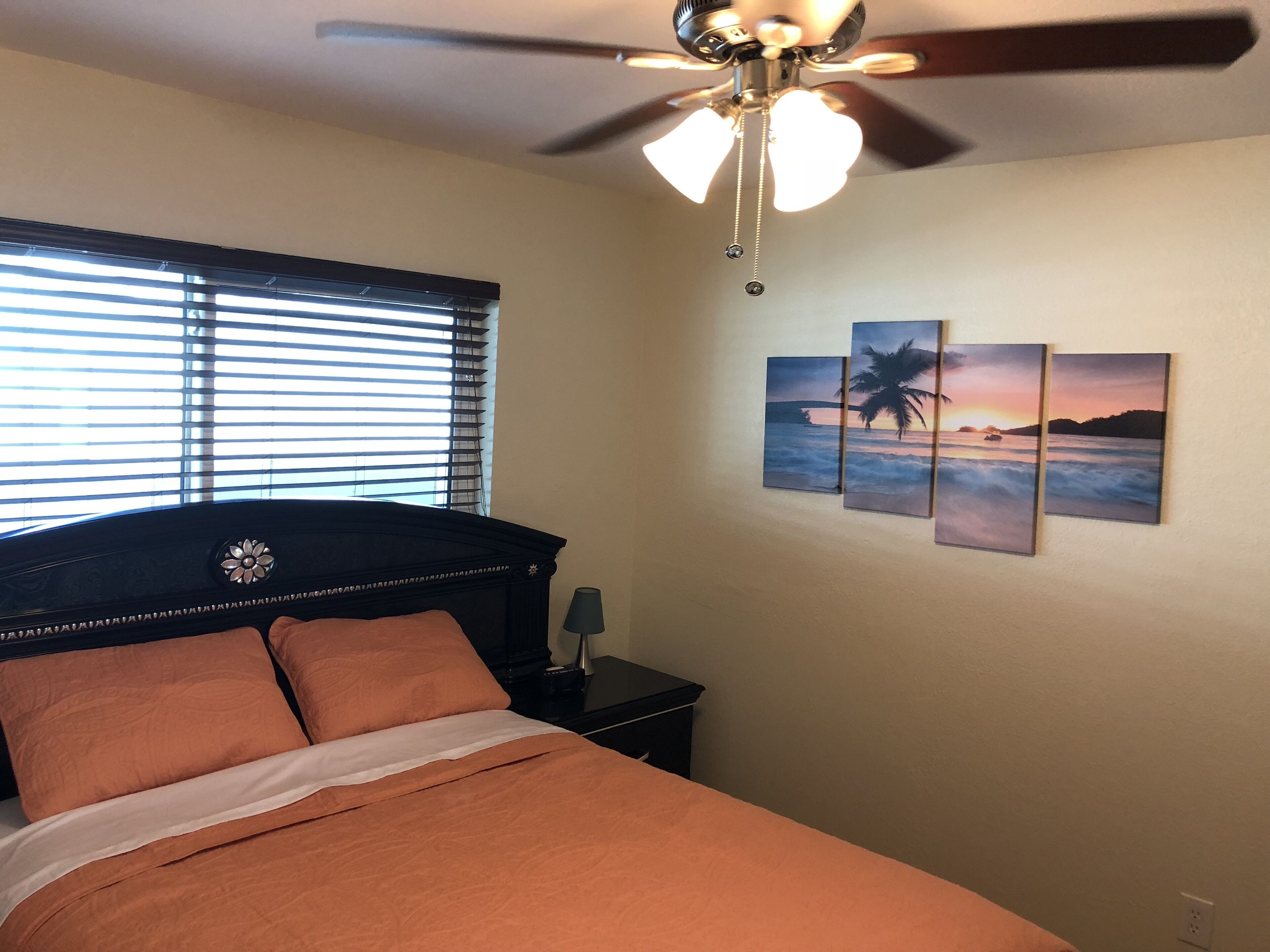 Ein Schlafzimmer mit einem Bett, einem Deckenventilator, einem Fenster mit Jalousien und drei an der Wand befestigten Bildern.