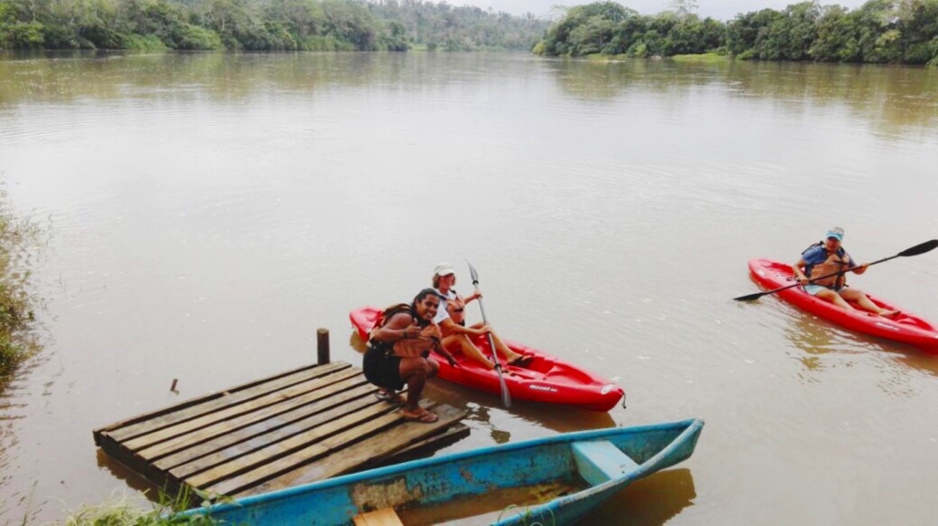 A calm river with two red kayaks and a blue canoe docked at a wooden pier.