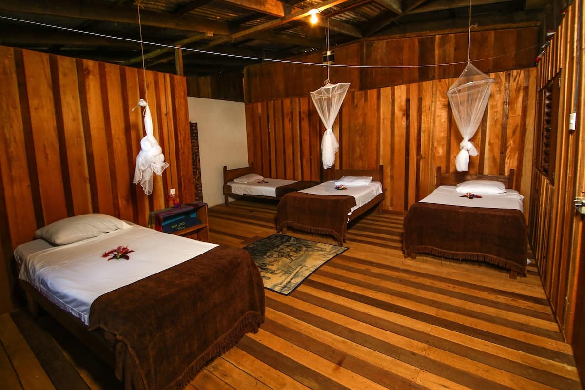 A room with wooden walls and floors, two beds with white linens, a wooden bench, and mosquito nets hanging from the ceiling.